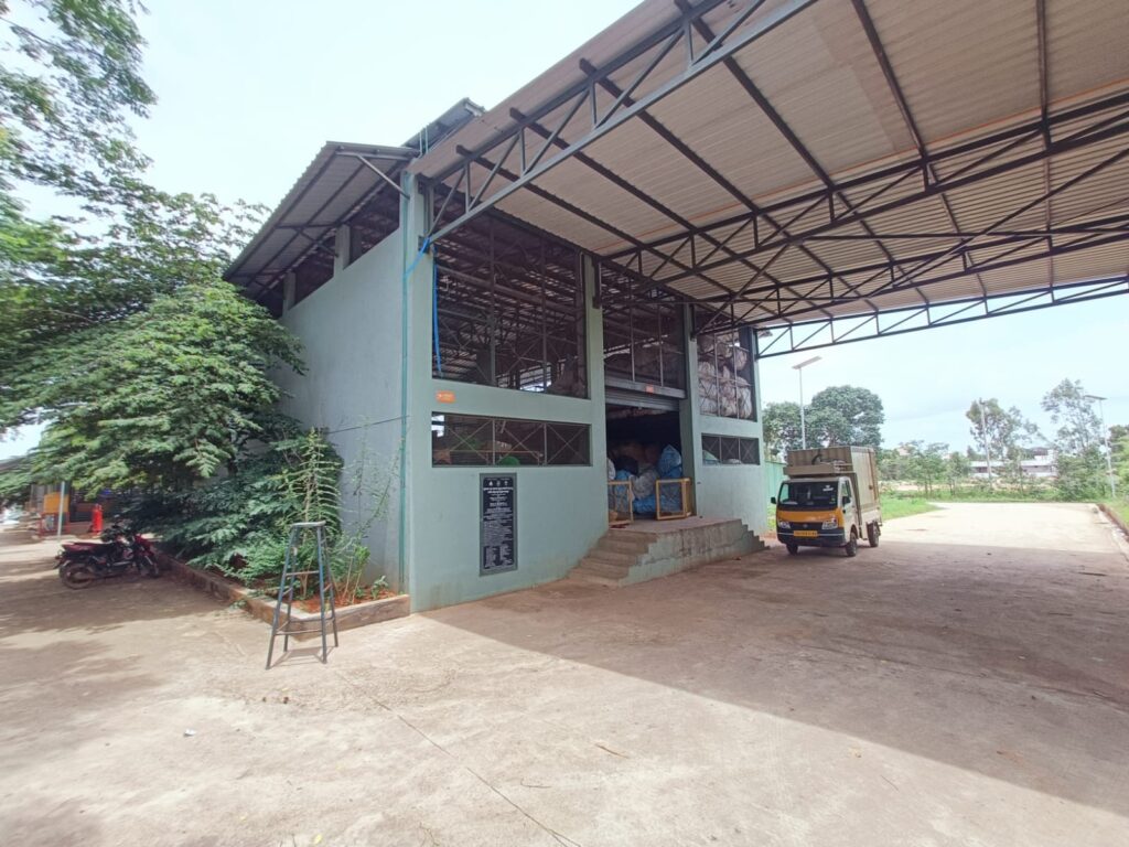 Exterior view of an EcoGram waste management facility in rural Bengaluru, with a covered loading bay and a collection truck parked outside.