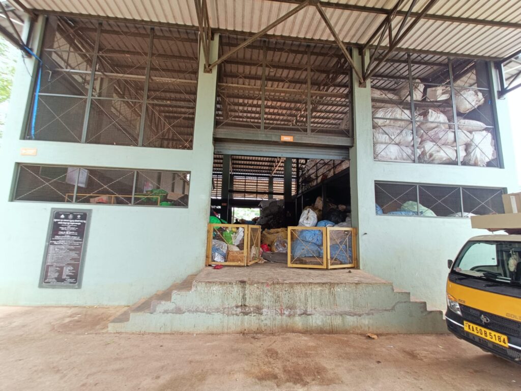 Entrance of a rural dry waste collection centre in Bettahalasuru, with sorted waste stored in mesh crates and sacks, and a collection vehicle parked to the side.