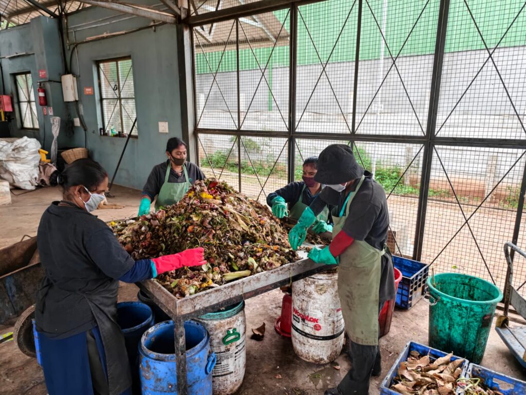 Four workers wearing masks, gloves, and aprons sort a large pile of organic food waste on a metal sorting table at an indoor composting facility, with collection bins and barrels arranged around them.